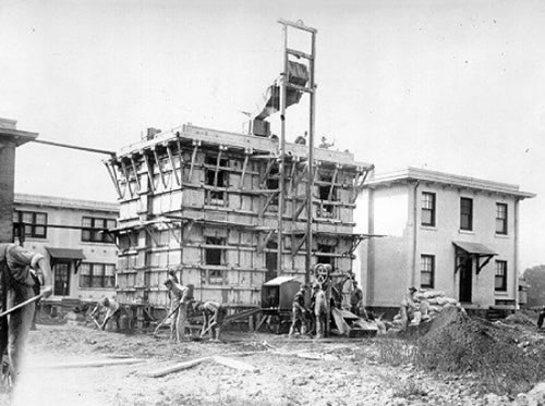 View of a concrete house being constructed. The building is flanked by scaffolding and small tower to pour concrete.
